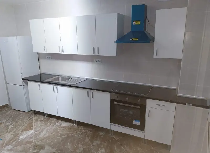 Cocina blanca en línea con encimera oscura, campana envuelta en plástico protector azul con su etiqueta de aviso, horno y vitrocerámica, alicatado gris y suelo porcelánico
