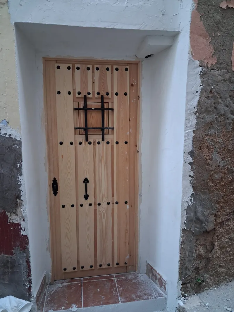 Puerta de calle de madera de pino con clavos negros y una reja de forja, en una fachada encalada