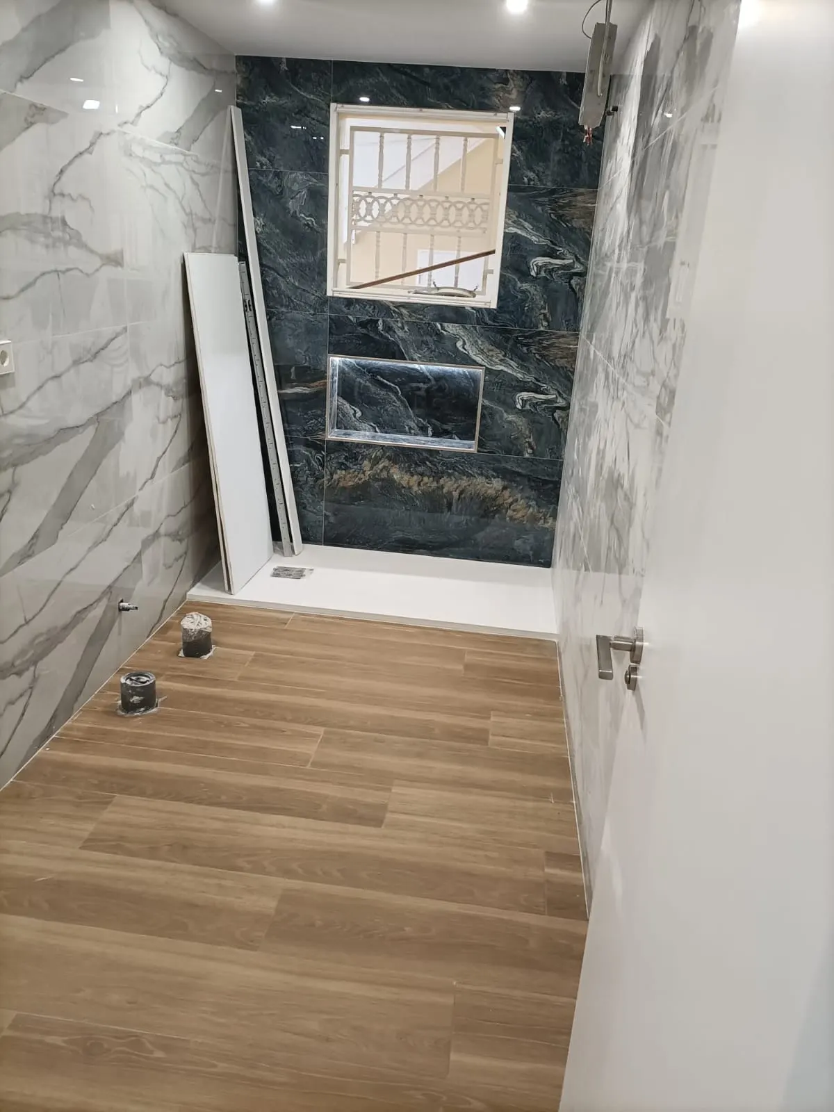 Baño alicatado en gran formato con mármol gris en un paño y piedra oscura veteada en el frente de la ducha, con hornacina, plato de ducha blanco colocado y suelo porcelánico imitación madera