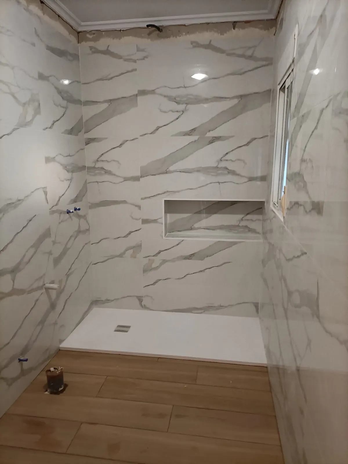 Zona de ducha alicatada en mármol blanco de gran formato con hornacina, plato de ducha blanco colocado y suelo porcelánico imitación madera