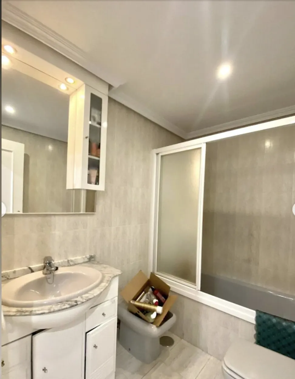 Baño con bañera y mampara corredera traslúcida, alicatado beige, mueble blanco con espejo sobre el lavabo y una caja de cartón junto al inodoro