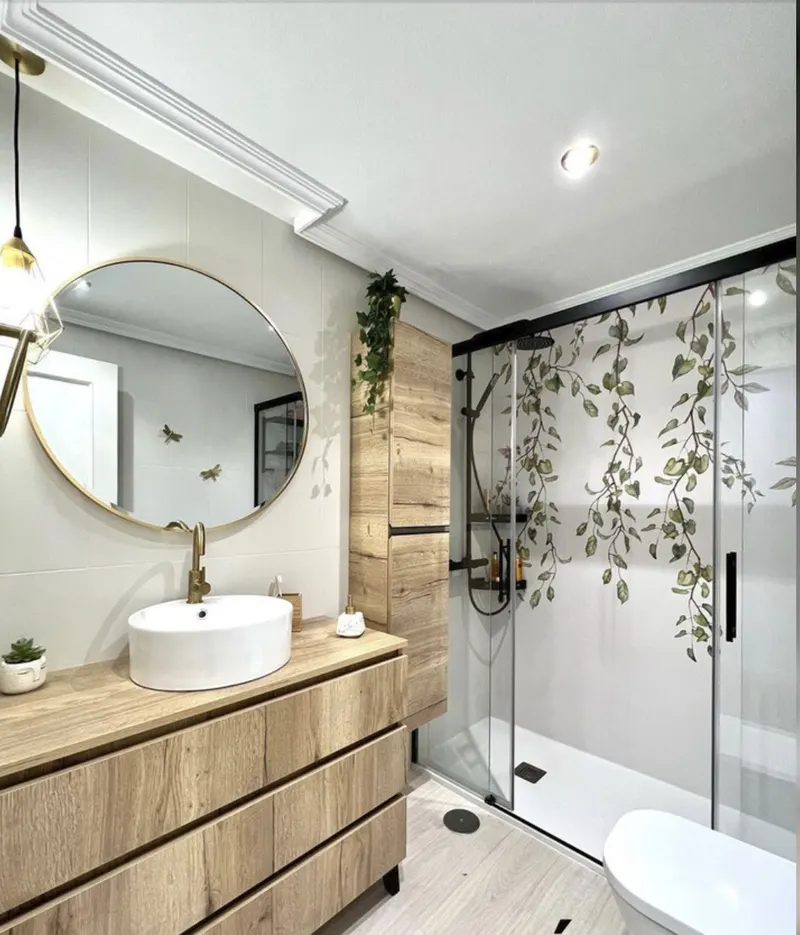 Baño con mueble de madera y lavabo sobre encimera, grifería dorada, espejo redondo y ducha con mampara de vidrio con estampado vegetal